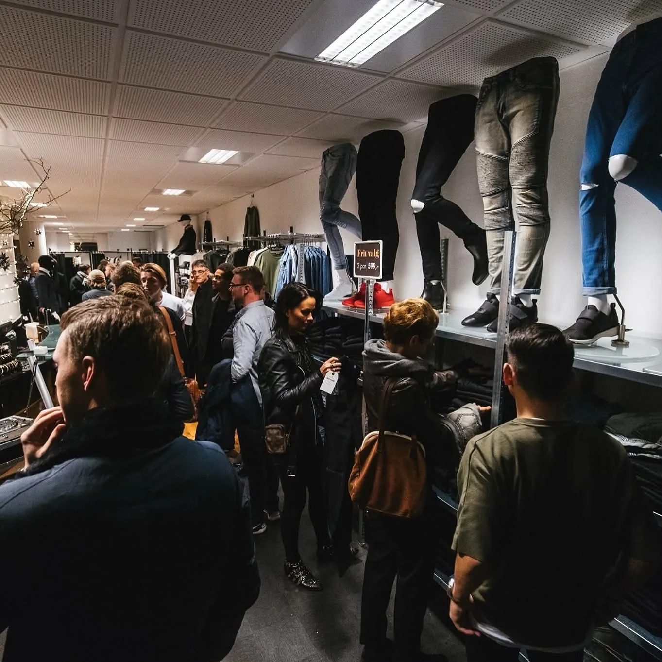 En fyldt tøjbutik med kunder, jeans på væggen og tilbudsskiltning.
